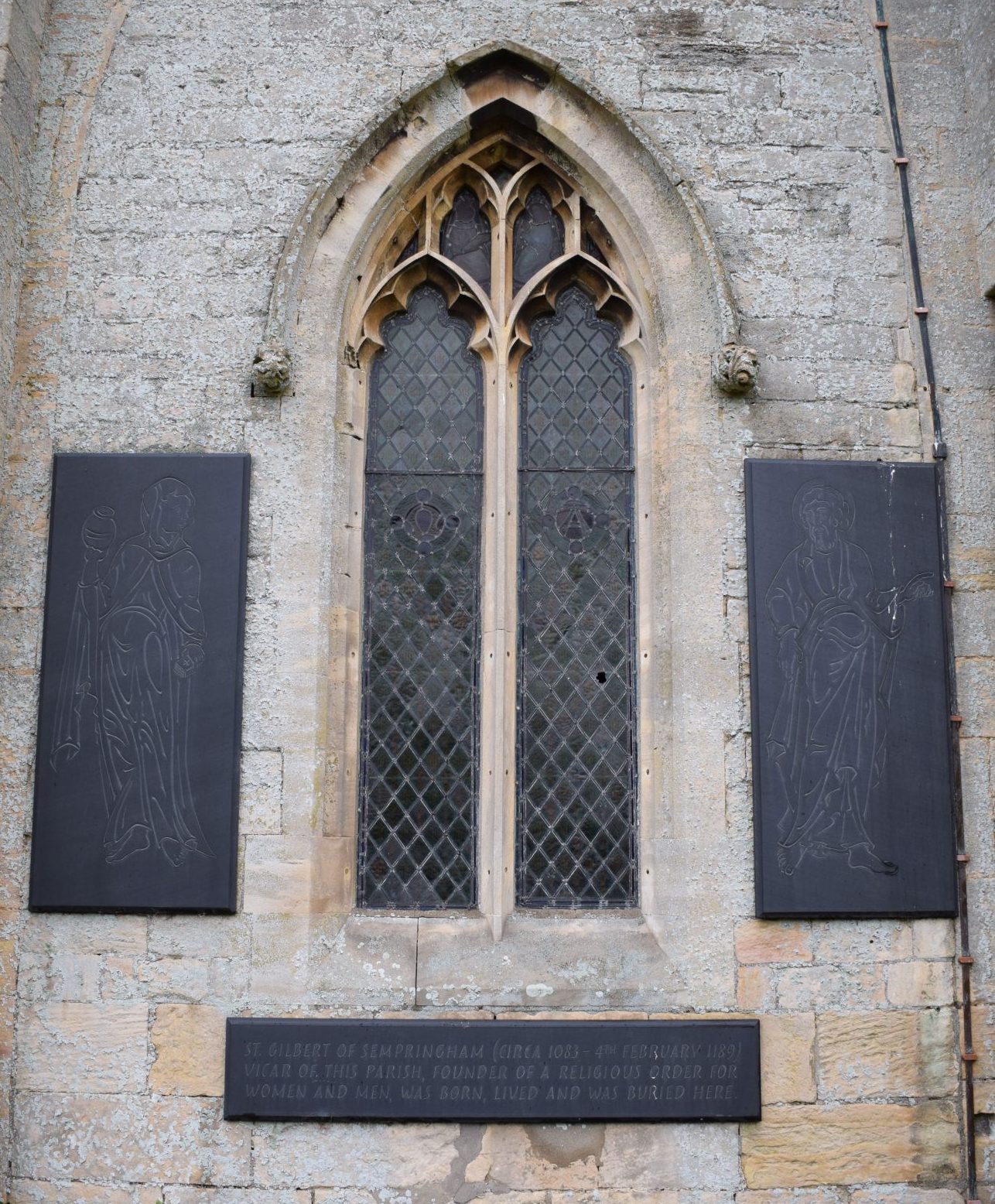 Church window surrounded by slate plaques on each side and below the window featuring images of a Gilbertine nun and cannon and a memorial to St Gilbert.