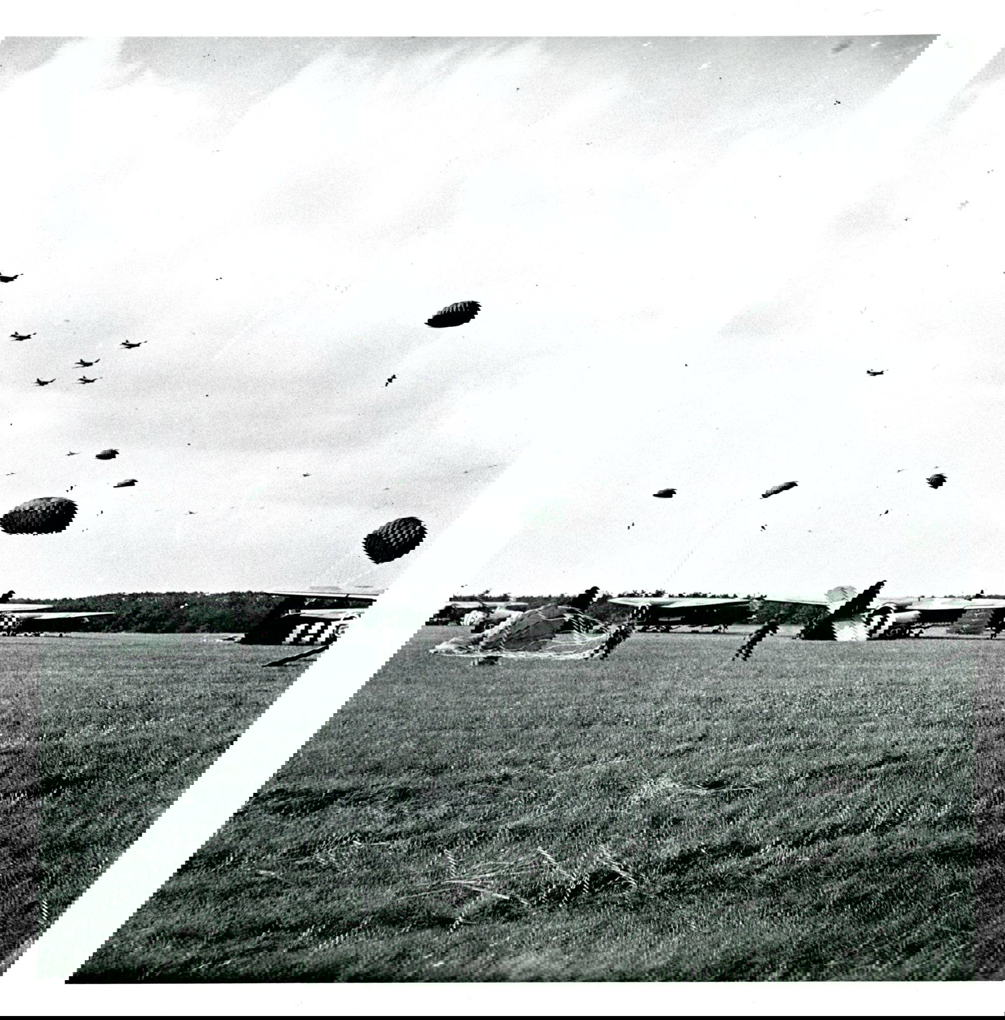 Paratroopers landing at Arnhem in September 1944 Parachutes descend from planes in the sky as soldiers land on an open field. Several aircraft are visible on the ground in the background.